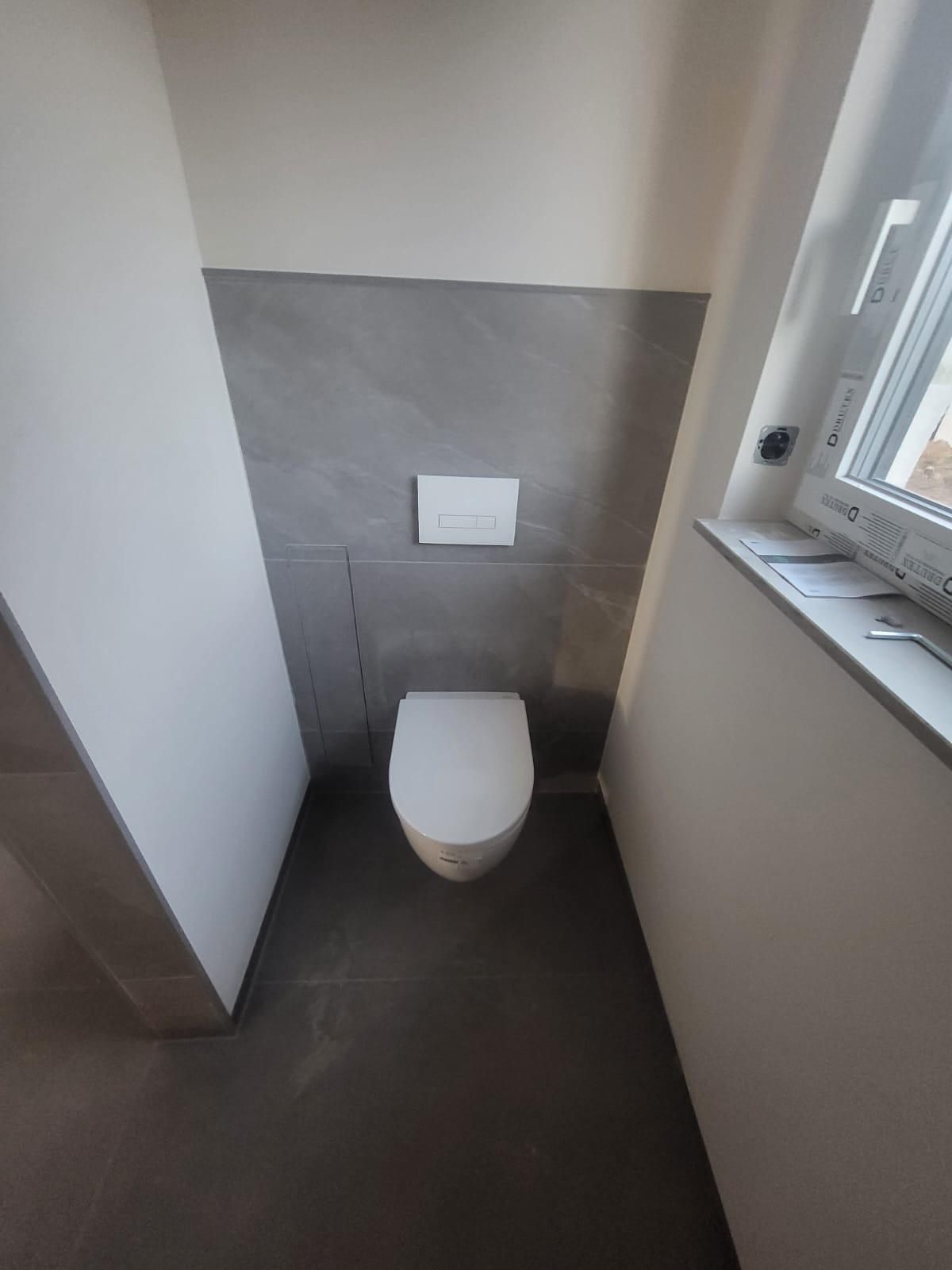 Fertig befliestes Toilettenelement mit Fussboden Wand-WC in einem modernen Bad mit grauen Fliesen und Fenster.